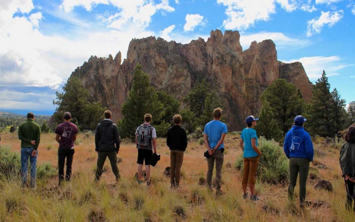 A group of people stand in a line, facing away from the camera. They appear to be looking at a rock formation in the distance.